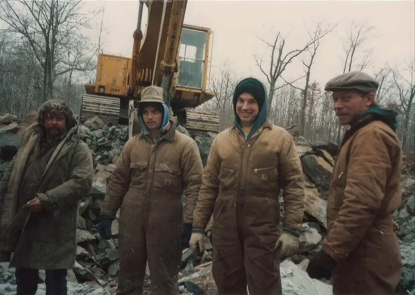 Crew of four in winter Carhartts standing in front of a CAT excavator on a snowy demolition site.