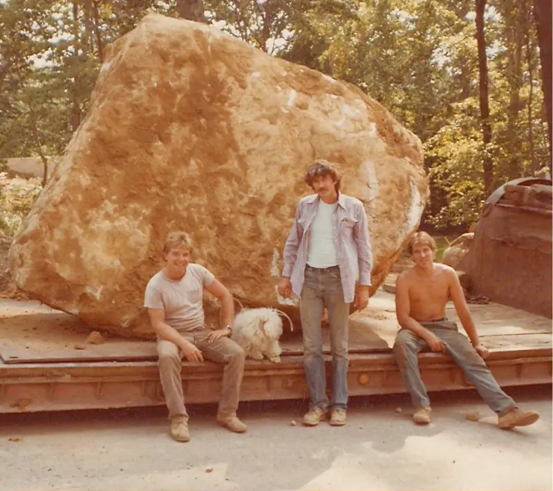 Crew posing on a flatbed with a large boulder, woods behind.