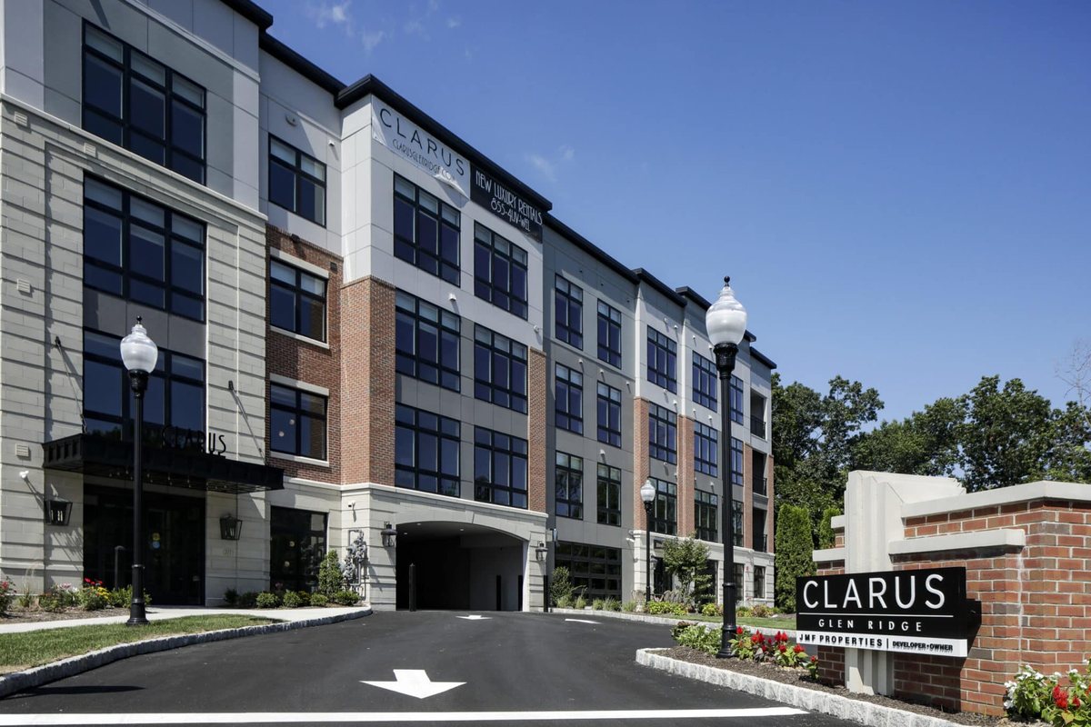 Front exterior of Clarus Glen Ridge with branded monument sign, dark brick base, light gray upper facade, paved drive and parking with directional arrow striping