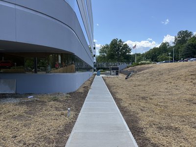 New concrete sidewalk along the base of the Marotta Controls building with freshly seeded, hay-covered slopes.