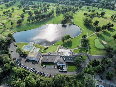 Wide aerial of the clubhouse, pond, and course with the newly paved parking lot along the tree line.