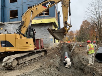 CAT 321D excavator lowering a large concrete pipe segment into an open utility trench while two workers in orange safety vests guide the placement