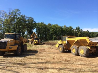 Two yellow articulated dump trucks and excavator clearing a wooded lot under blue sky