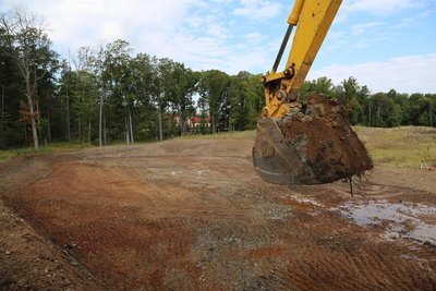 Excavator bucket dumps wet soil onto graded earth pad near tree line