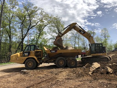 Cat excavator loads soil into Cat 725C articulated truck on a wooded lot