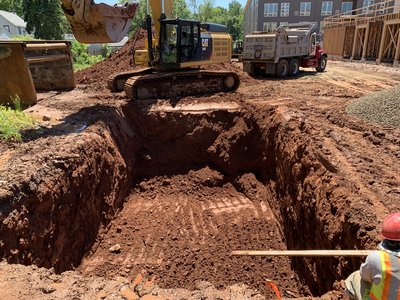Cat excavator digging a deep red-clay foundation pit beside a tandem dump truck
