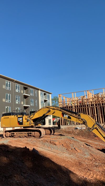 Cat excavator on a graded pad beside a wood-framed multistory building under construction