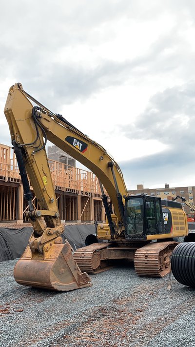 Cat excavator parked on gravel beside framed multistory buildings under overcast sky