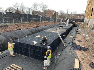 Workers assemble plastic stormwater chamber crates inside geotextile-lined excavation under gray sky
