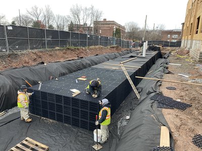 Crew constructs modular plastic infiltration chambers within fenced excavation lined with black geotextile