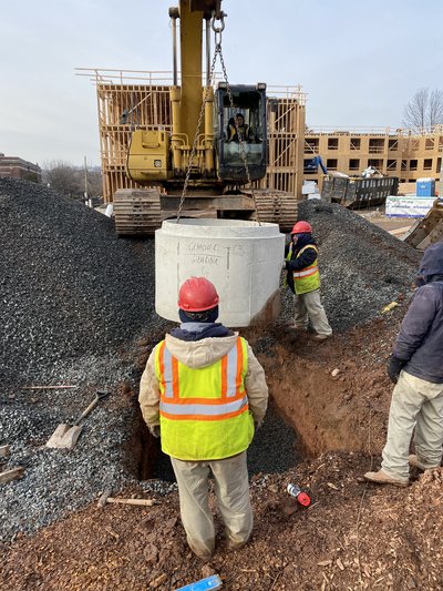 Excavator lowers precast concrete drainage structure as workers in vests guide placement into excavation
