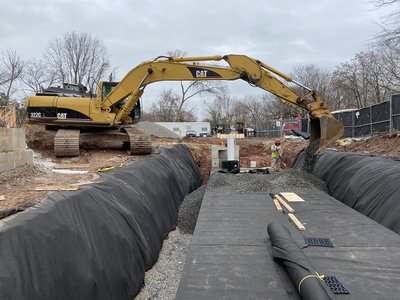 CAT 322C excavator places gravel envelope along geotextile-lined stormwater trench with worker observing