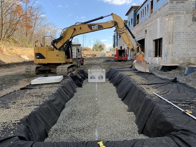 CAT excavator beside gravel-lined stormwater trench with geotextile fabric and precast inlet structure