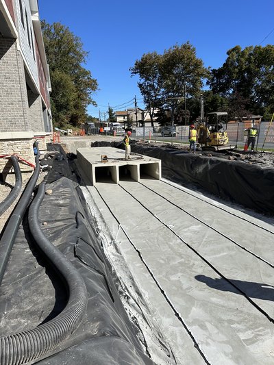 Workers install precast concrete detention vault beside building with excavator and liner geotextile