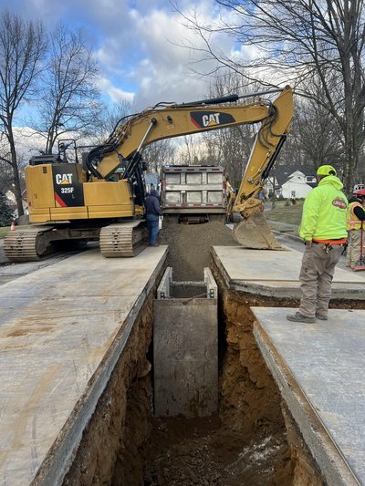 CAT 325F excavator places concrete drainage structure into open trench on residential street
