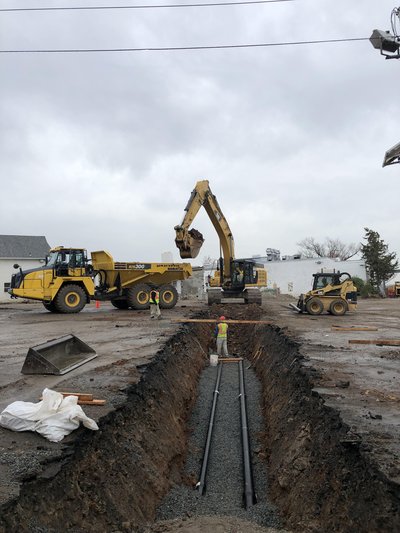 Deep utility trench with twin pipes on stone bedding, excavator and articulated dump truck beyond
