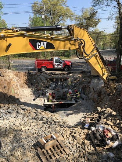 Excavator and crew work in rocky pit installing utility structure beside flatbed truck