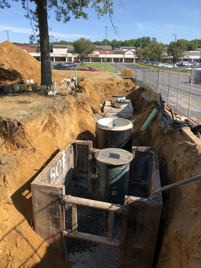 Deep trench with shoring box and stacked manhole sections being set beside parking lot