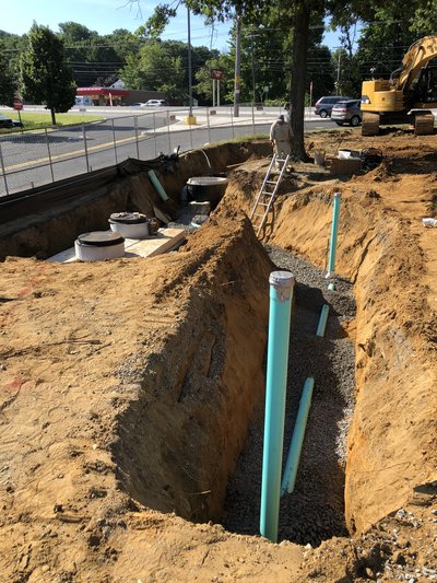 Open utility trench with vertical green pipe risers on stone bedding beside roadway