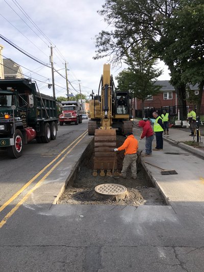 Crew works in road cut around manhole frame with excavator and dump trucks staged