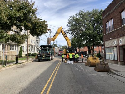 Excavator and dump trucks block residential street while crew works at sandbagged road cut