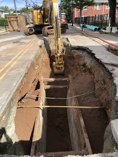 Deep road-cut trench with shoring box and exposed pipe, excavator parked at edge