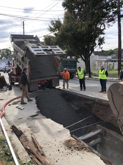 Dump truck discharges asphalt into open road cut while crew supervises backfill operation