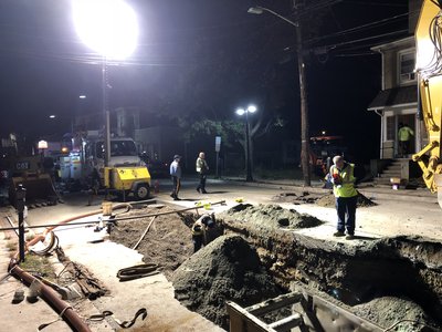 Nighttime utility excavation lit by floodlights with workers, compressor, and excavator on residential street