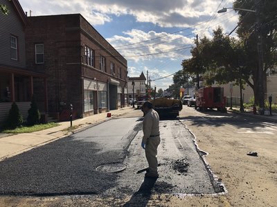 Worker inspects fresh asphalt patch over manhole repair on sunny urban street with traffic