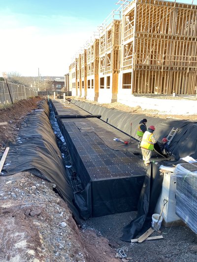 Tarped trench excavation alongside wood-framed building under construction with workers in safety vests