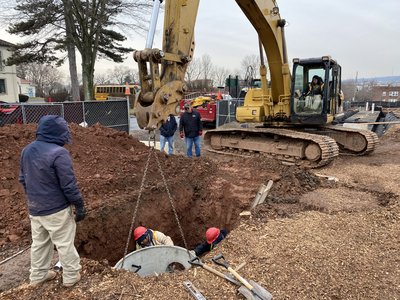 Excavator lowers concrete manhole structure into pit while two workers guide placement below