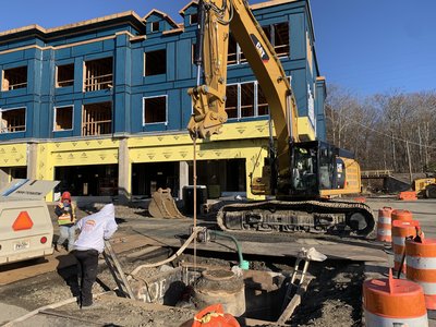 Excavator works over open utility pit beside framed multi-story building under construction
