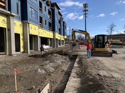 Mini excavator backfills narrow utility trench along curb beside multi-story building under construction
