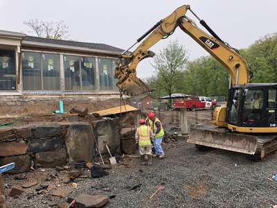Excavator lifts a large stone block as workers rebuild a fieldstone retaining wall