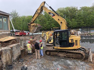 Two excavators work alongside crew members rebuilding a stone retaining wall on rainy day