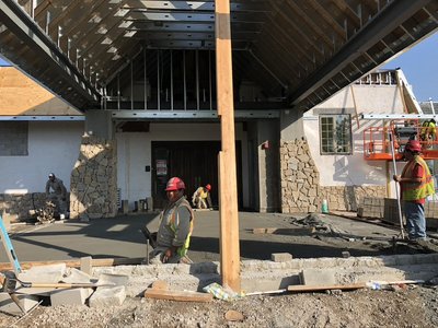 Workers finish a freshly poured concrete walkway beneath a timber-framed building entrance
