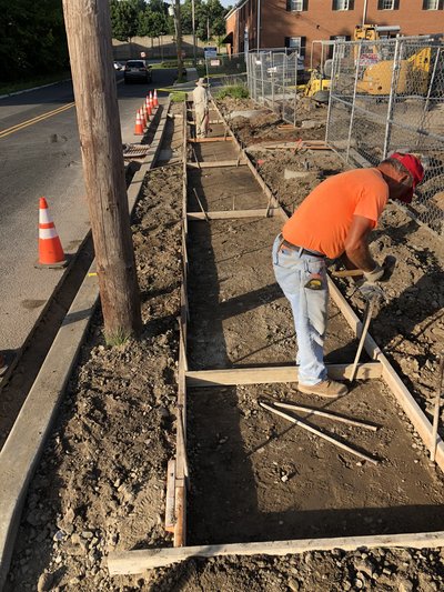 Worker frames sidewalk forms along roadside with traffic cones and chain-link fencing behind