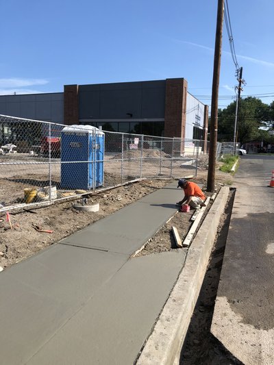 Worker finishes wet concrete sidewalk along curb edge beside fenced commercial building
