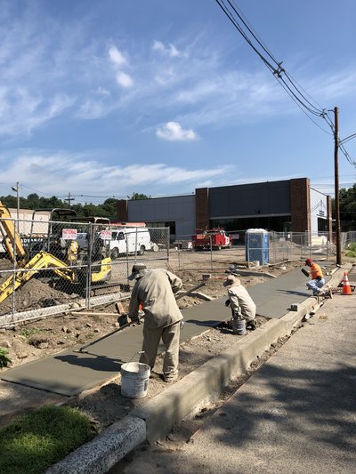 Three workers finish concrete sidewalk pour along curb with excavator behind chain-link fence
