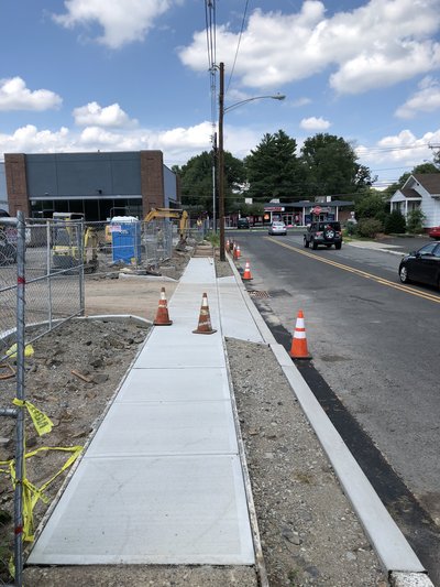 Completed concrete sidewalk runs alongside roadway with traffic cones marking edge protection
