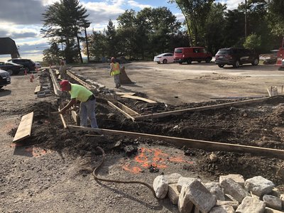 Worker sets cobblestone curbing in formed trench with stacked granite blocks staged nearby