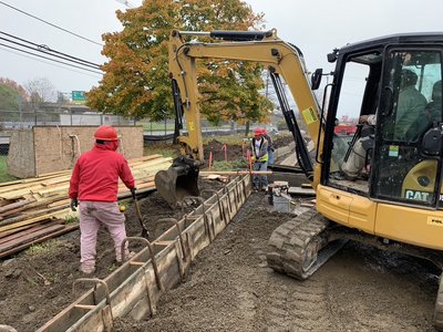 Excavator grades soil beside formwork while a worker rakes material along the wall