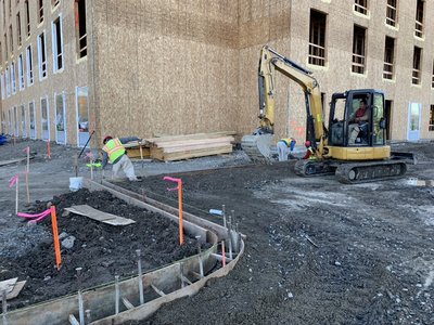Worker prepares curved formwork for a sidewalk pour beside a wood-sheathed building