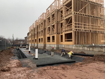 Crew installs gravel drainage along foundation wall beside a wood-framed multistory building