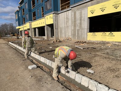 Two workers set granite cobblestone curbing in fresh concrete beside a multistory building