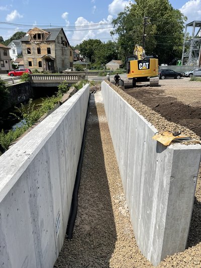 Tall poured concrete retaining wall channel with excavator working alongside roadway bridge