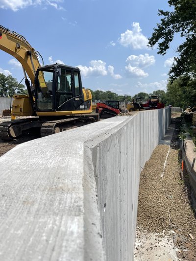 Excavator parked beside cured curved board-form concrete retaining wall on summer day