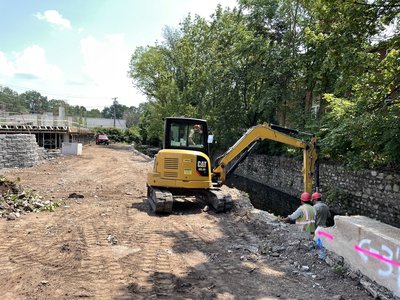 Mini excavator working stone channel as workers in vests inspect retaining wall base