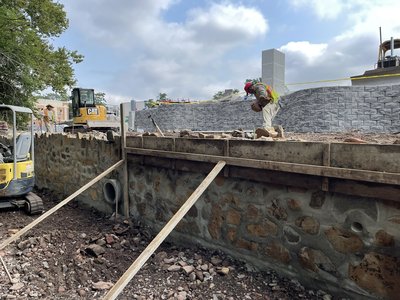 Mason setting capstones atop fieldstone-faced concrete retaining wall with tracked loader nearby