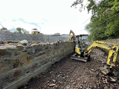 Mini excavator working rocky channel beside completed fieldstone-veneered concrete retaining wall